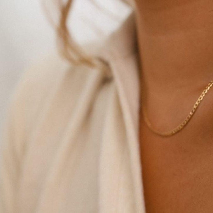 A close-up of a person wearing a light-colored, textured shirt slightly open at the collar, showcasing their neck and chest adorned with the elegant Marie Chain 18k gold-filled necklace by LLUME Jewelry.