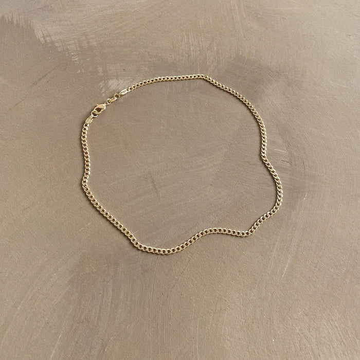 The Marie Chain by LLUME Jewelry, a thin 18k gold-filled necklace with a simple clasp, is displayed in a loose oval shape on a smooth, light brown surface.