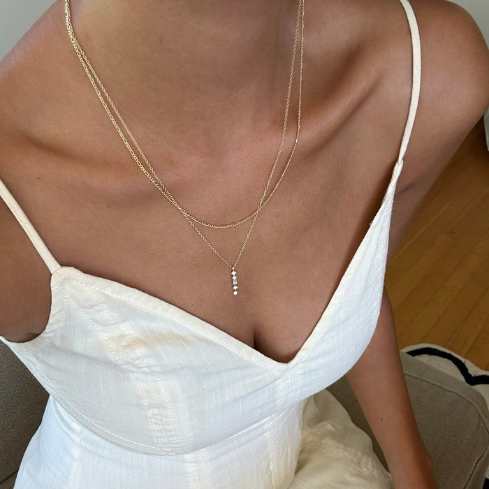 Seated indoors on a light chair with wooden floors beneath, a woman in a white spaghetti strap dress wears LLUME Jewelry's Midnight Daily Set—a delicate necklace set with small pendants, including one adorned with tiny pearls.