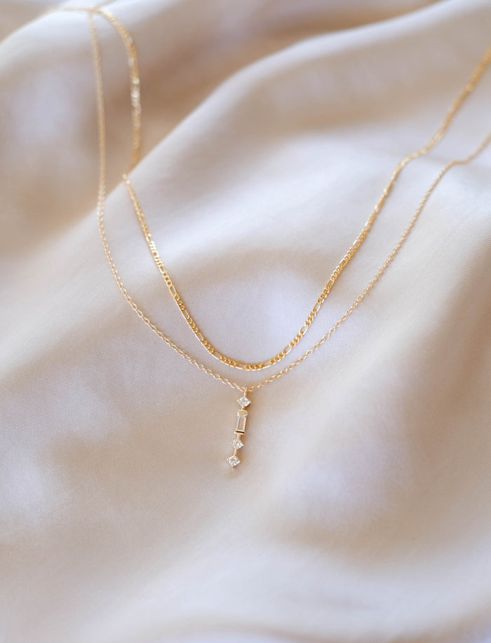 The Midnight Daily Set by LLUME Jewelry features two delicate gold necklaces—one plain and one with a pendant of three small geometric gemstones—styled on an ivory fabric background.