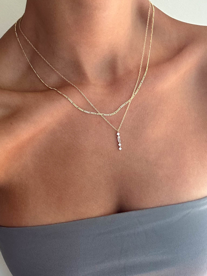 A person wears a strapless grey top and the LLUME Jewelry Midnight Daily Set, featuring a delicate necklace with a small vertical pendant, against a neutral background.