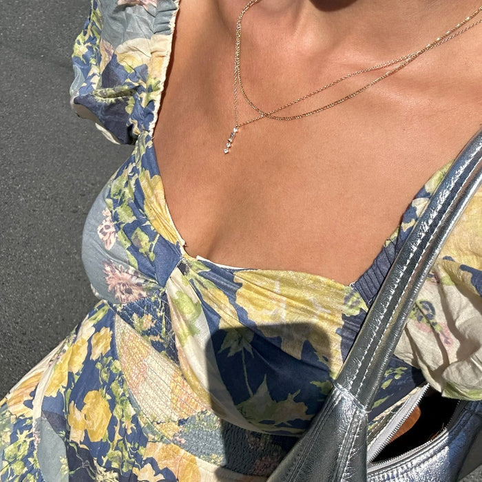 A close-up of a person in a floral dress with blue, yellow, and green hues. They wear LLUME Jewelry's Midnight Daily Set and carry a shiny silver bag over their shoulder as sunlight highlights their skin and outfit.