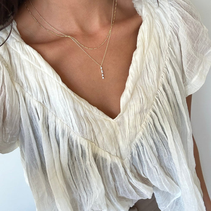 A person wears the LLUME Jewelry Midnight Daily Set—a delicate necklace featuring small beads—paired with a white, flowy, textured blouse with a deep V-neckline against a neutral background.