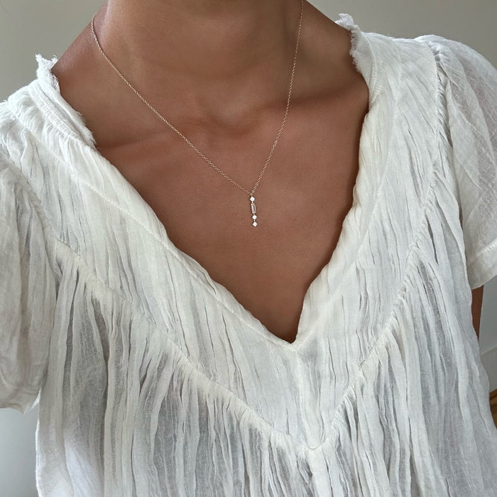A woman wears a textured white V-neck blouse and the LLUME Jewelry Midnight Necklace, a dainty gold-plated chain with small pendants. The image is cropped to highlight her neck and upper chest.