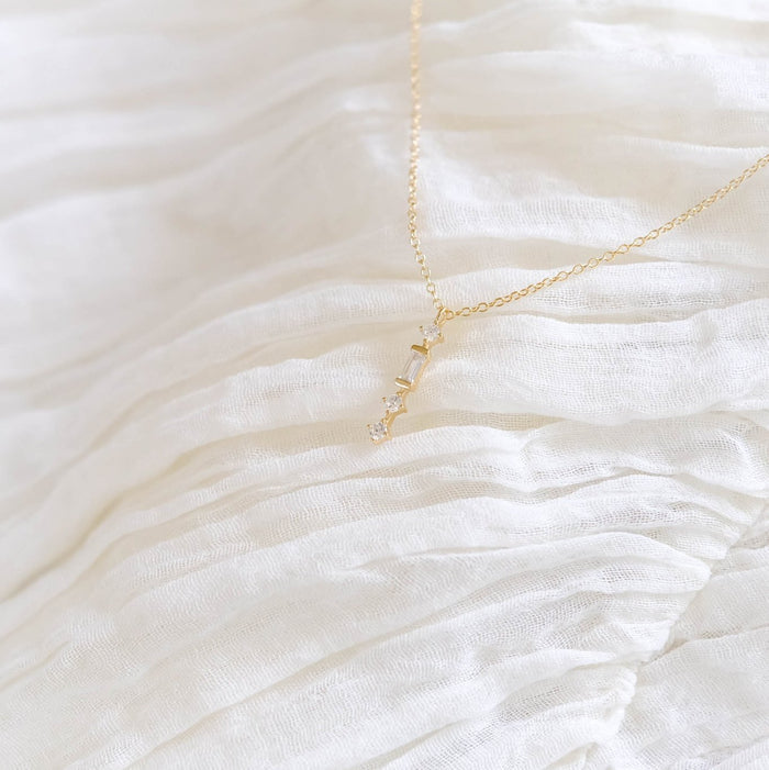 The Midnight Necklace by LLUME Jewelry, a delicate gold-plated piece with a LOVE pendant, is displayed on textured white pleated fabric.