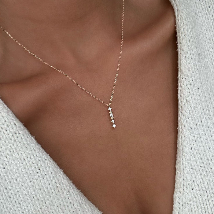 The Midnight Necklace by LLUME Jewelry, a gold plated and dainty piece with a vertical row of sparkling gems, is worn elegantly against the collarbone with a white textured V-neck sweater.