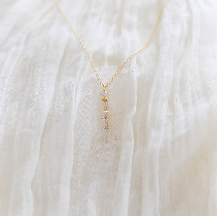 The Midnight Necklace by LLUME Jewelry is a dainty gold-plated piece with a vertical diamond-accented pendant, elegantly showcased on soft white textured fabric.