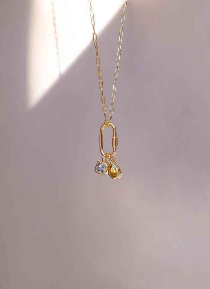 A delicate gold chain necklace by LLUME Jewelry features two small square charms—one clear, one yellow—suspended from the Mini Charm Carabiner, photographed against a soft, light beige background.