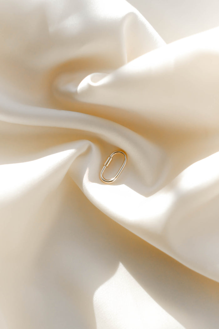 The Mini Charm Carabiner by LLUME Jewelry, a small gold accessory, lies on creamy white satin, creating an elegant and minimal scene with gentle light and smooth textures.