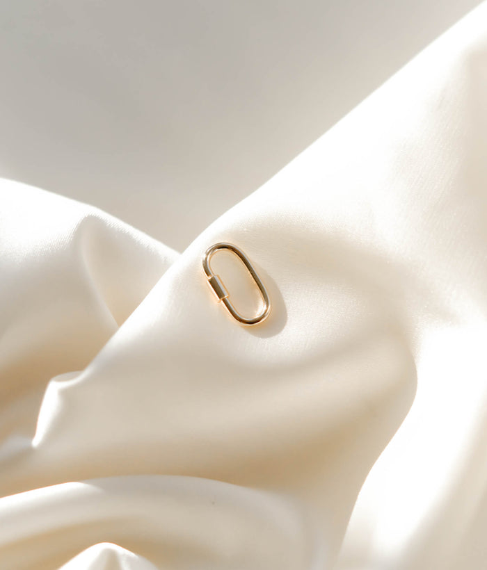 The LLUME Jewelry Mini Charm Carabiner is displayed on soft, cream-colored satin with gentle folds, highlighting its elegant and minimalistic design.
