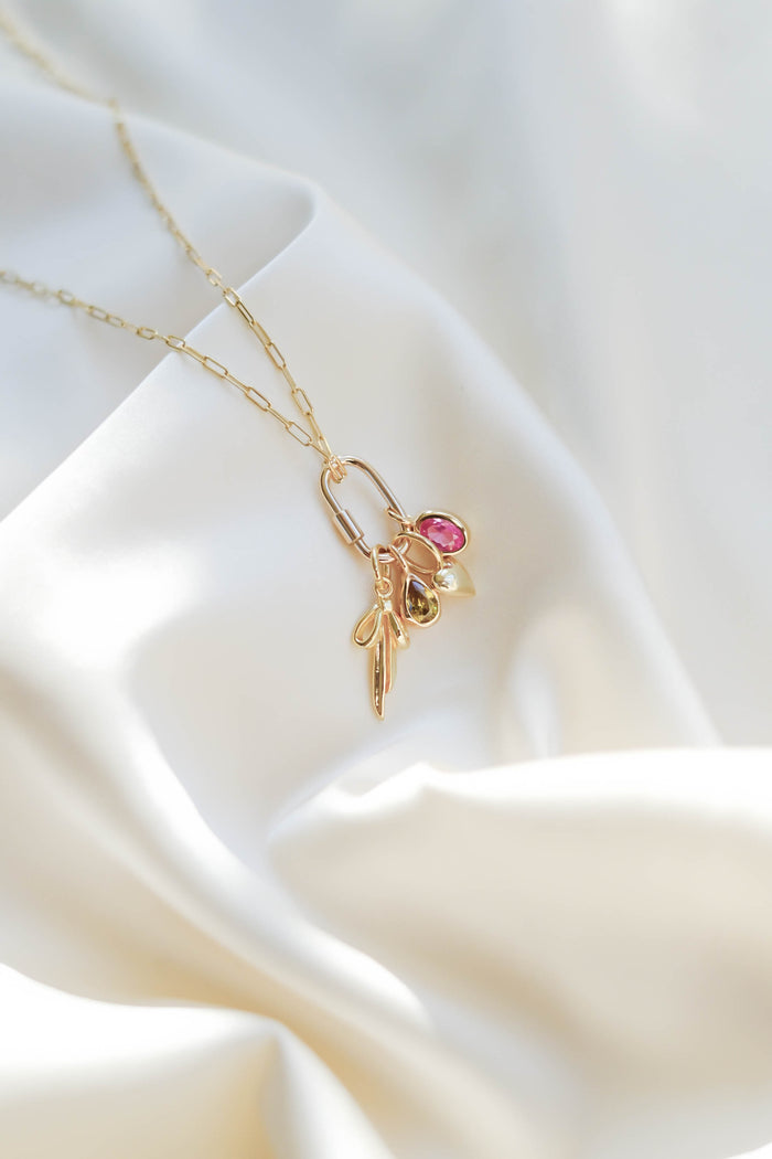 The LLUME Jewelry Mini Charm Carabiner, a delicate gold accessory with three small charms—a pink gemstone, a pearl, and a leaf—rests on soft white satin-like fabric in natural light.