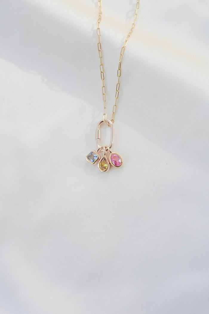The LLUME Jewelry Mini Charm Carabiner gold chain necklace, featuring three gemstone charms—a Mini Carabiner Charm, square blue stone, and oval pink stone—rests on a white background, making it an ideal accessory for any occasion.