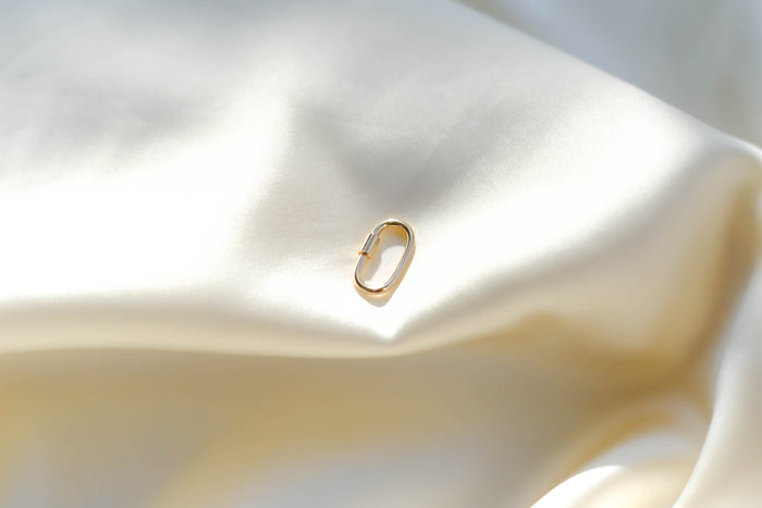 The LLUME Jewelry Mini Charm Carabiner is a small gold clasp displayed on smooth white satin, reflecting soft light—an ideal accessory to complement your favorite necklace.