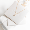 The Moment Necklace by LLUME Jewelry features a delicate gold star pendant, displayed on an off-white embossed card atop a fabric-wrapped box with dried grass accents—an elegant accessory for any occasion.