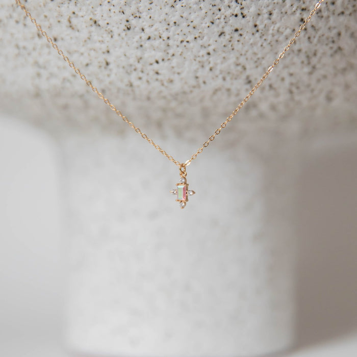 The LLUME Jewelry Moment Necklace is a dainty gold piece with a vertical baguette gemstone pendant, shown on textured white ceramic. This elegant necklace brings subtle charm to any look.