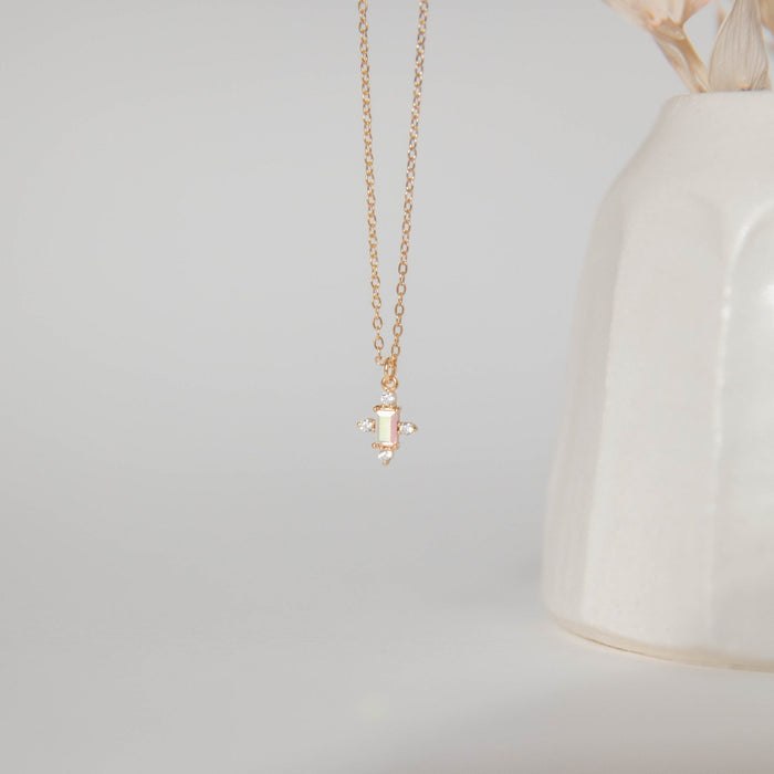 The Moment Necklace by LLUME Jewelry features a delicate gold chain and a small rectangle pendant with tiny gemstones, shown hanging before a cream ceramic vase on a light background.