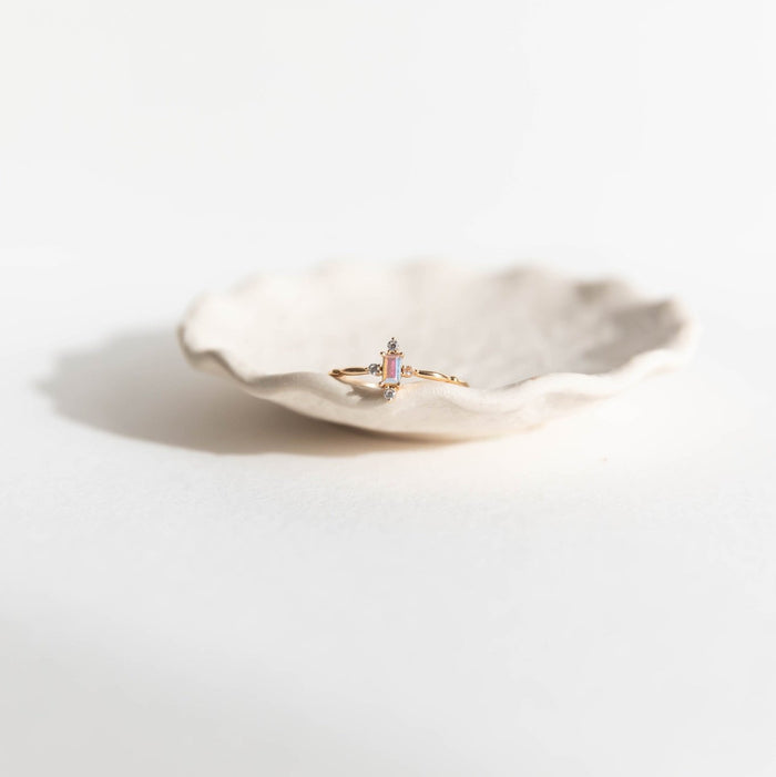 The Moment Ring by LLUME Jewelry, a delicate 14k gold-filled band with a small gemstone, is shown in a white scalloped ceramic dish on a softly lit white surface.