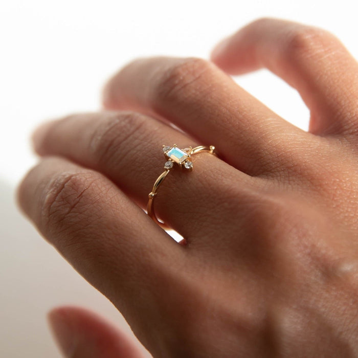 A close-up of a hand wearing LLUME Jewelry's Moment Ring, a delicate 14k gold-filled band with a central oval opal and tiny clear side stones, set against a soft, neutral background.