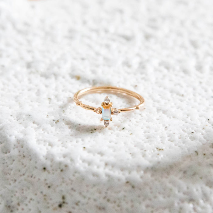 The LLUME Jewelry Moment Ring is a delicate 14k gold-filled band with a vertical opal and four tiny diamonds, shown on a textured white surface—an elegant, timeless piece.