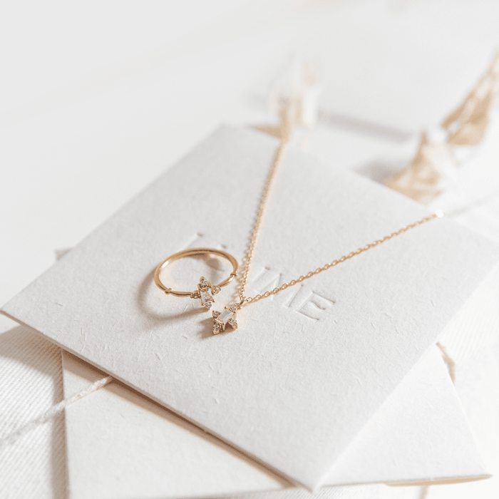 The LLUME Jewelry Moment Set, featuring a ring and necklace with small cross-shaped pendants adorned with clear stones, is displayed on a textured white card with embossed text against a softly lit, minimalistic background.