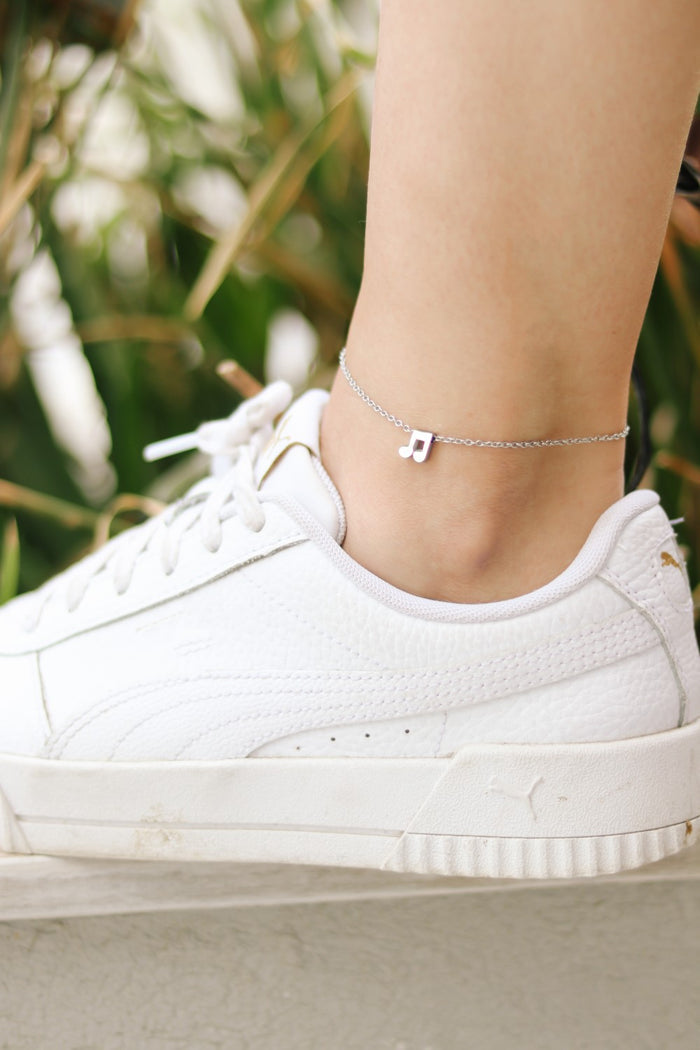 A silver chain anklet with a tiny music note charm worn on a person's ankle, paired with a white sneaker.