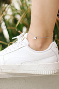 A silver chain anklet with a tiny music note charm worn on a person's ankle, paired with a white sneaker.