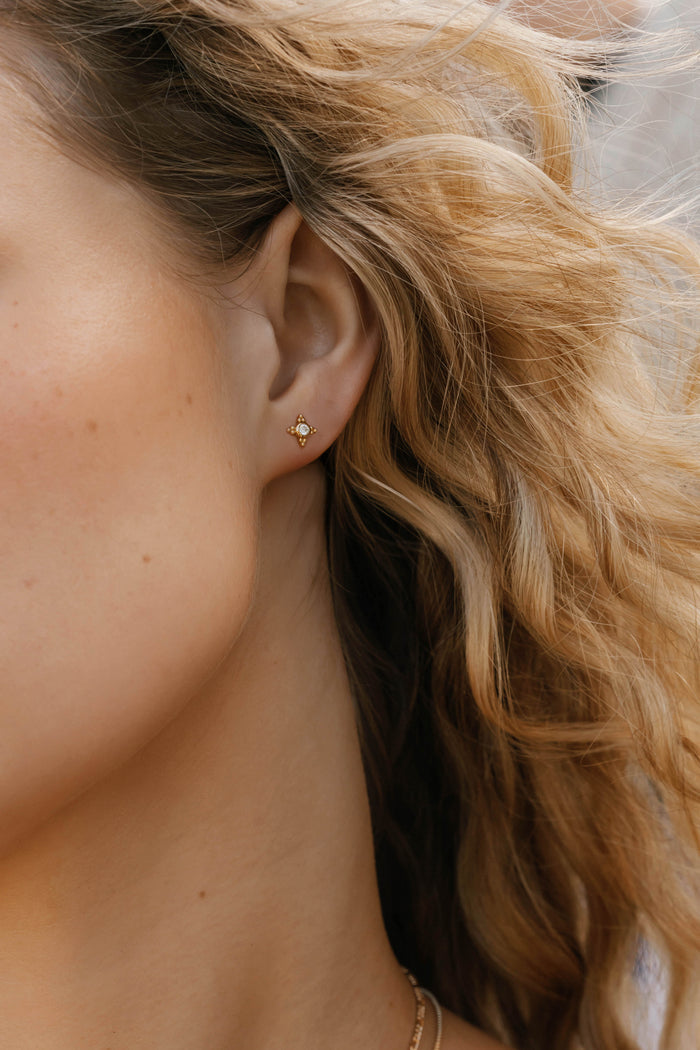 A close-up shows a woman's ear wearing LLUME Jewelry's Myra Studs—delicate earrings with a tiny central gemstone. Her wavy blonde hair frames her naturally toned skin, while a softly blurred background accentuates the jewelry.