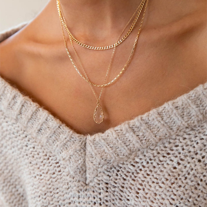 A close-up of a person's neck wearing LLUME Jewelry's New Era Bundle—a layered gold necklace set with a teardrop pendant—styled over a beige off-shoulder sweater, perfect for elevating your winter wardrobe.