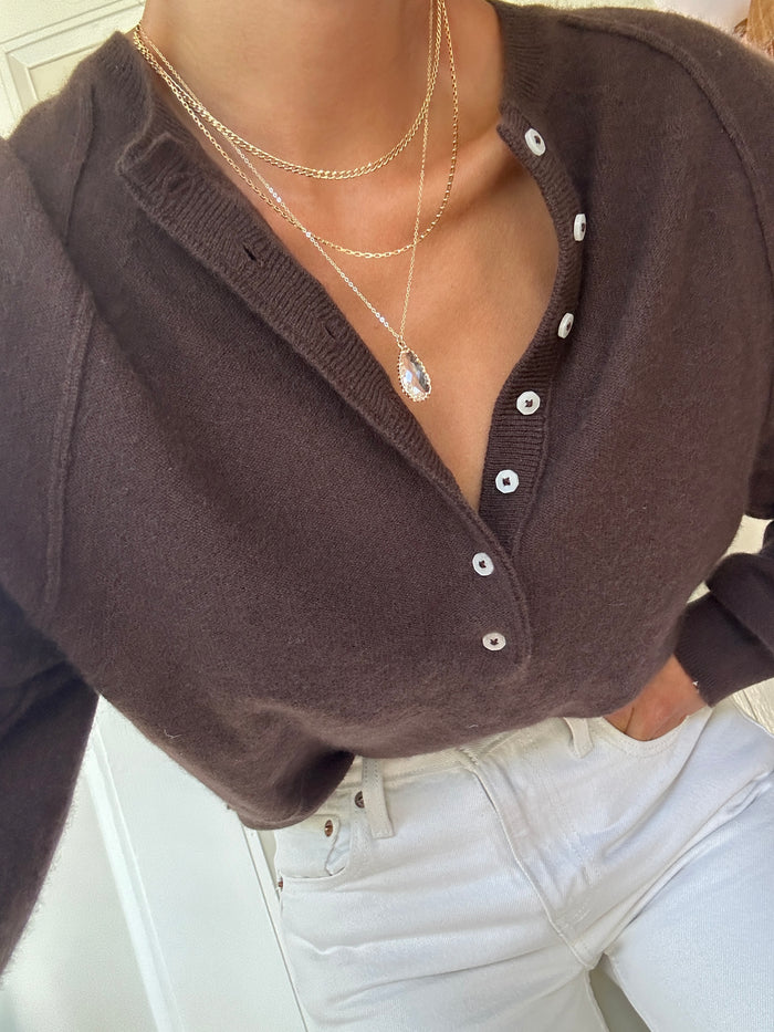A person in a dark brown buttoned sweater and white pants wears LLUME Jewelry's New Era Bundle layered necklaces, one with a teardrop-shaped pendant. The cropped image highlights chic winter style inspiration.