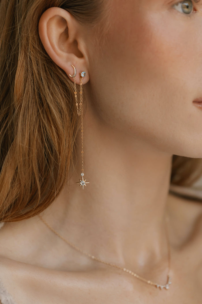 A close-up of a woman’s ear and neck features LLUME Jewelry's Night Sky Earrings—beautiful gold pieces with crescent moon and star details—layered elegantly, ideal for winter style.