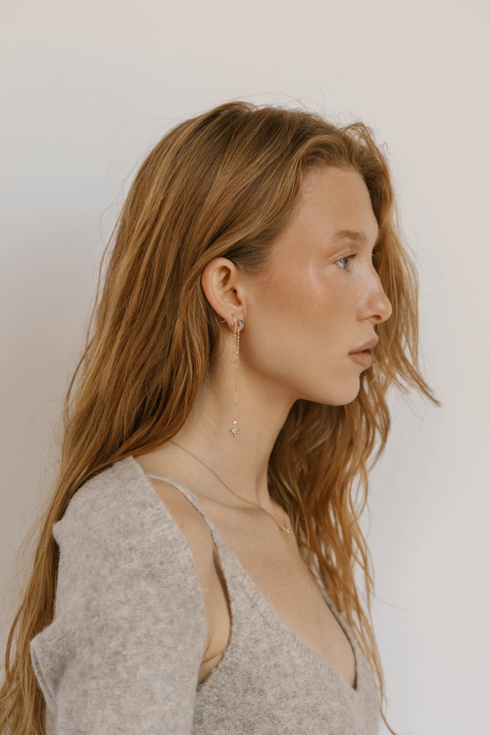 A young woman with long, wavy, light brown hair wears a light gray sweater, delicate jewelry, and LLUME Jewelry's Night Sky Earrings—a perfect choice for elegant winter style.