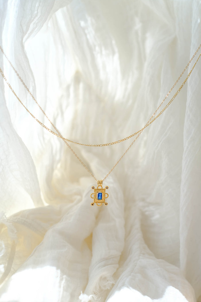 The Nina Daily Set by LLUME Jewelry features a delicate gold necklace with a turtle-shaped pendant and a sapphire blue Nina gemstone, beautifully displayed on soft, white textured fabric under gentle natural light.