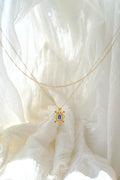 The Nina Daily Set by LLUME Jewelry features a delicate gold necklace with a turtle-shaped pendant and a sapphire blue Nina gemstone, beautifully displayed on soft, white textured fabric under gentle natural light.