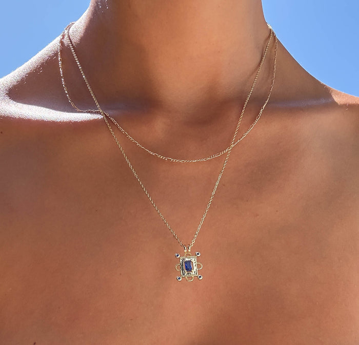 A person wears the LLUME Jewelry Nina Daily Set: a simple chain and a Sapphire Blue pendant, both gleaming against sunlit skin with a clear blue sky in the background.