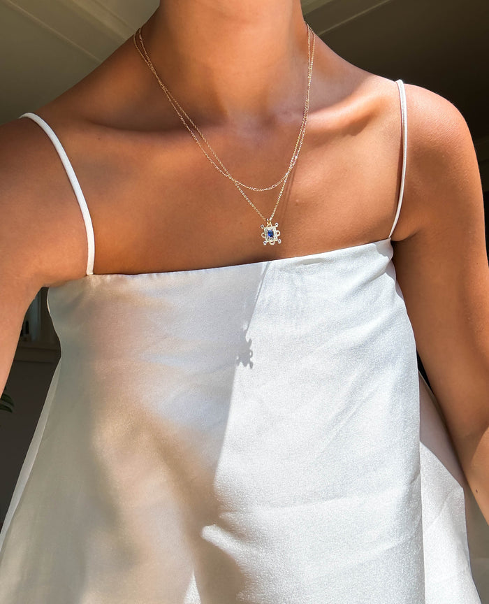 A person in a white satin spaghetti-strap top layers gold necklaces, including the LLUME Jewelry Nina Daily Set, and stands in sunlight.