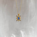 The Nina Necklace by LLUME Jewelry features a gold vermeil chain with a rectangular sapphire blue charm and small round accents, displayed against a soft, light-colored fabric background.