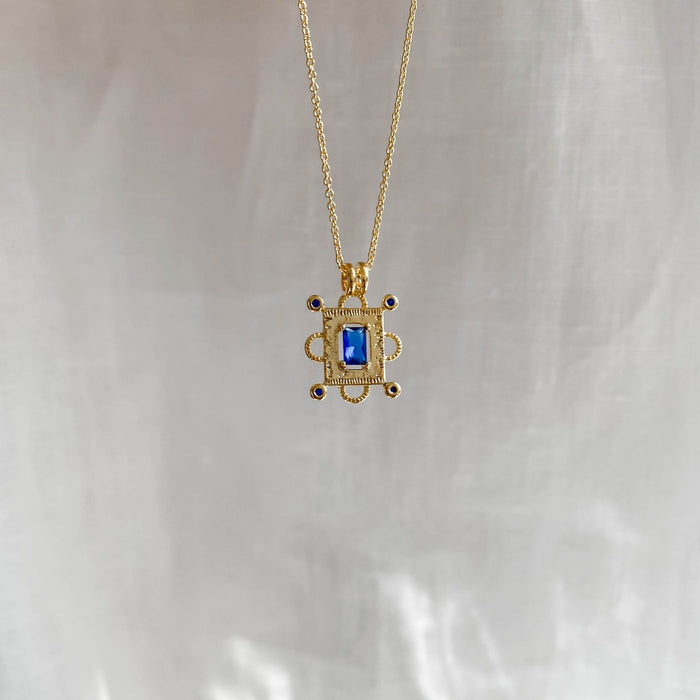 The Nina Necklace by LLUME Jewelry features a gold vermeil chain with a rectangular sapphire blue charm and small round accents, displayed against a soft, light-colored fabric background.