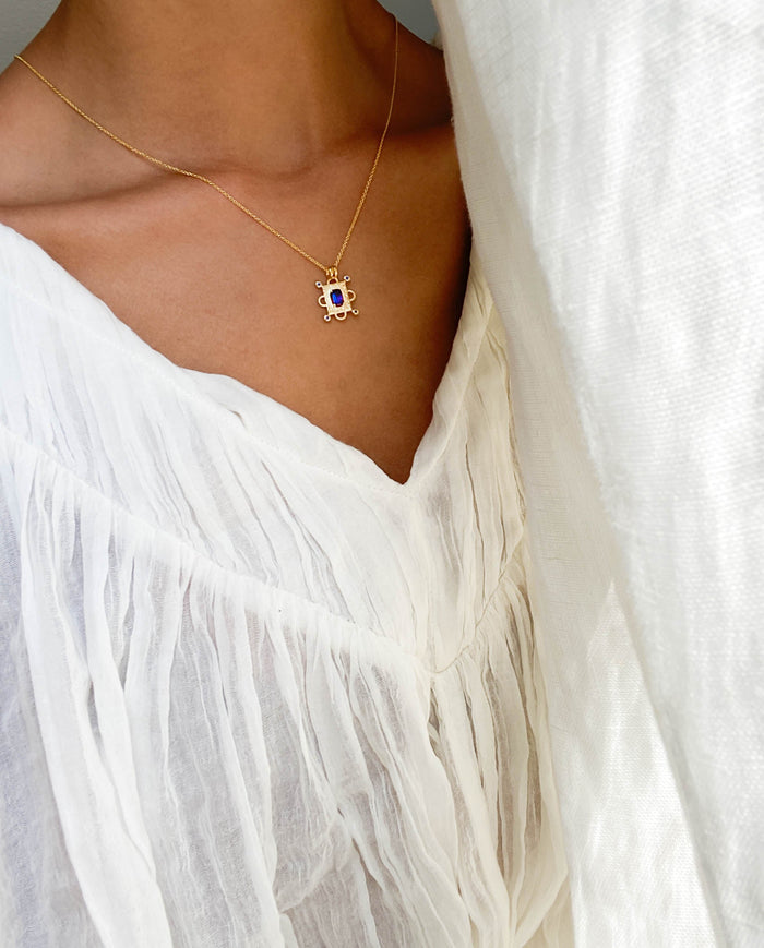 A person wearing a sheer white textured blouse with a deep V-neckline and the Nina Necklace by LLUME Jewelry— a delicate gold plated vermeil piece adorned with purple and white stones.