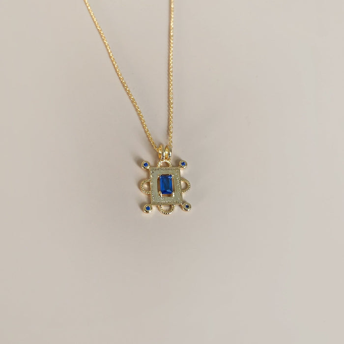 The Nina Necklace by LLUME Jewelry is a gold-plated vermeil piece with a turtle-shaped pendant, featuring a sapphire blue charm at the center and smaller blue stones on each limb, shown against a plain light background.