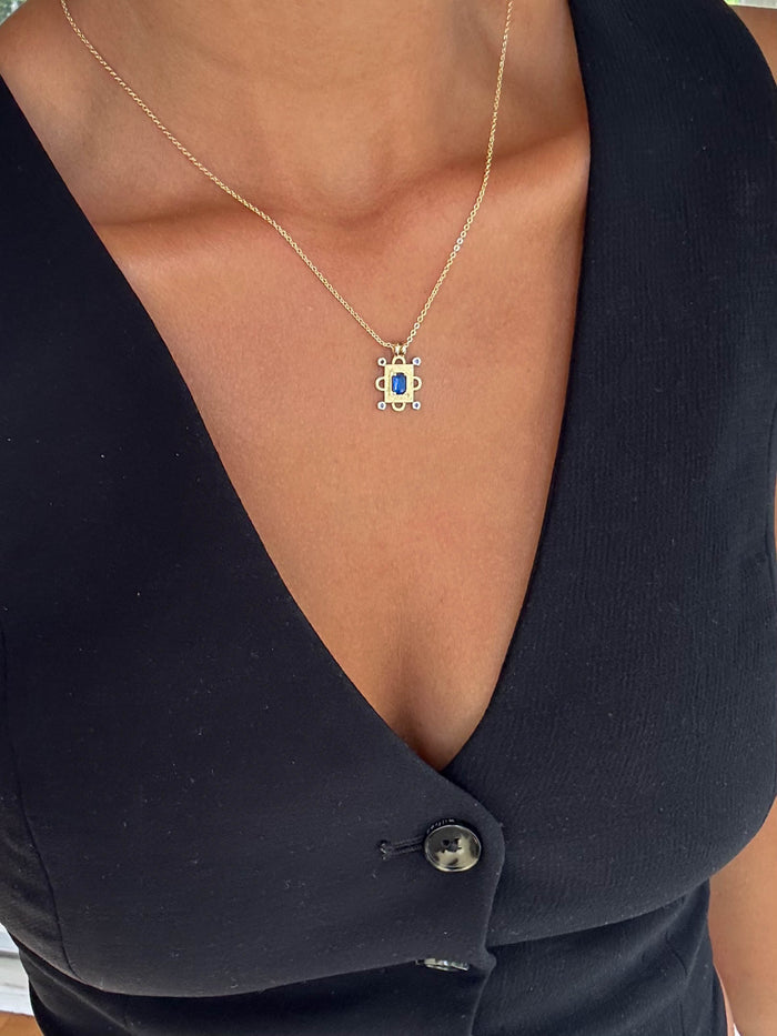 A person in a deep V-neck black top with button detail showcases the LLUME Jewelry Nina Necklace—a gold pendant necklace with a rectangular sapphire blue charm and white stones.