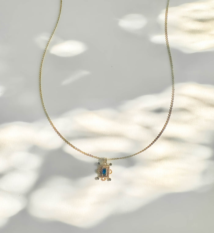 The Nina Necklace by LLUME Jewelry features a gold pendant with a rectangular sapphire blue charm and small round accents, shown on a white surface with soft, dappled light and shadow patterns.