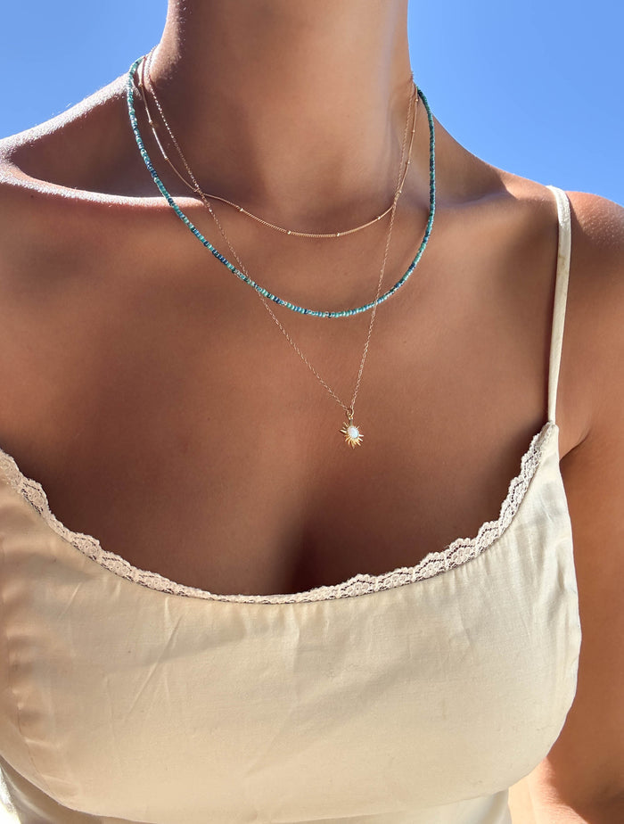 A person wearing a cream lace-trimmed camisole and the LLUME Jewelry Olandria Set—three layered necklaces with turquoise beads, gold chains, and a sun pendant—stands against a clear blue sky, perfect for any jewelry collection.