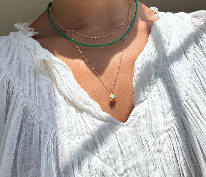 A person wears the Olandria Set by LLUME Jewelry: a green choker, thin gold chain, and gold pendant with gemstone, layered over a textured white V-neck blouse—an elegant jewelry statement.