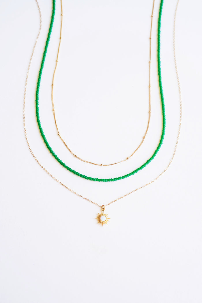 The Olandria Set by LLUME Jewelry features three layered necklaces: green beads, a gold sun-shaped pendant chain, and a delicate gold chain, beautifully displayed on a white background.