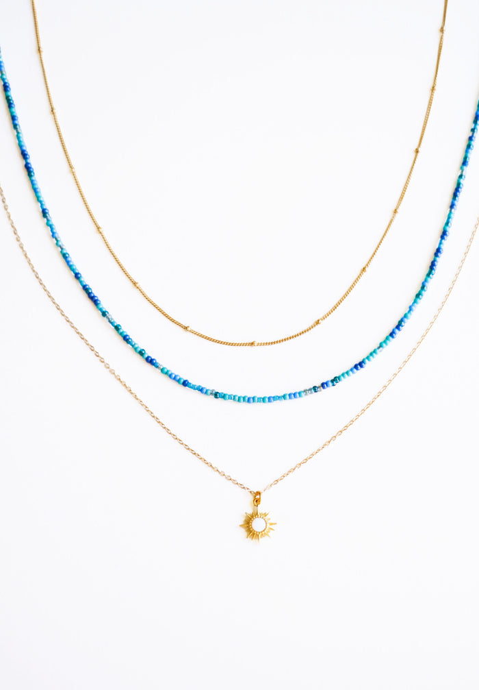 The Olandria Set by LLUME Jewelry features three layered necklaces—a gold chain, blue beaded strand, and gold sun pendant—arranged on a white background to bring versatile style to any outfit.