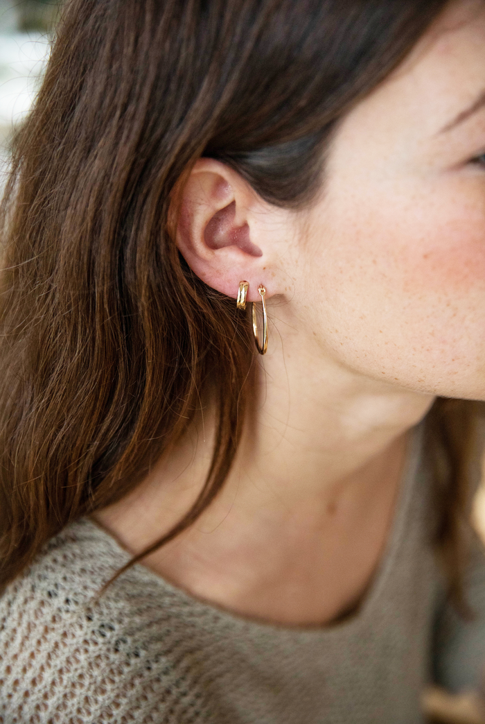 Close-up of a woman’s ear with long brown hair, wearing the elegant On my Way Stack earring set by LLUME Jewelry and a beige knit top. Her fair skin and natural look are softly illuminated by golden light.