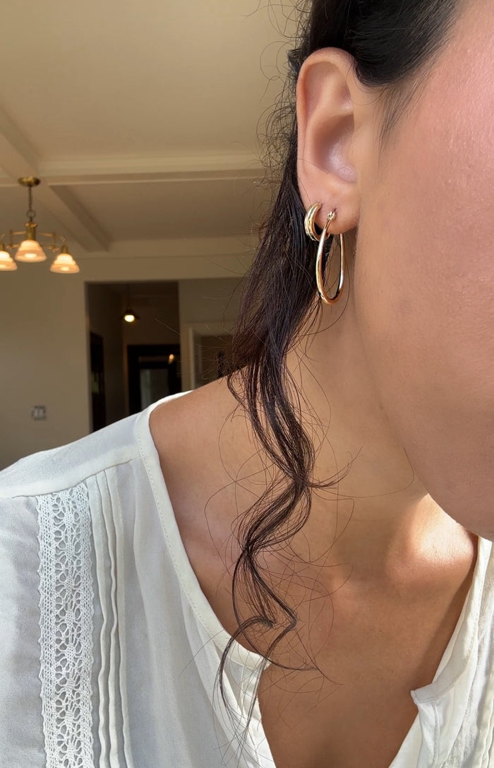 Close-up of a woman in a white lace blouse, her ear adorned with LLUME Jewelry's On my Way Stack. A loose wavy strand of dark hair frames her face, softly illuminated by warm indoor lighting in the background.