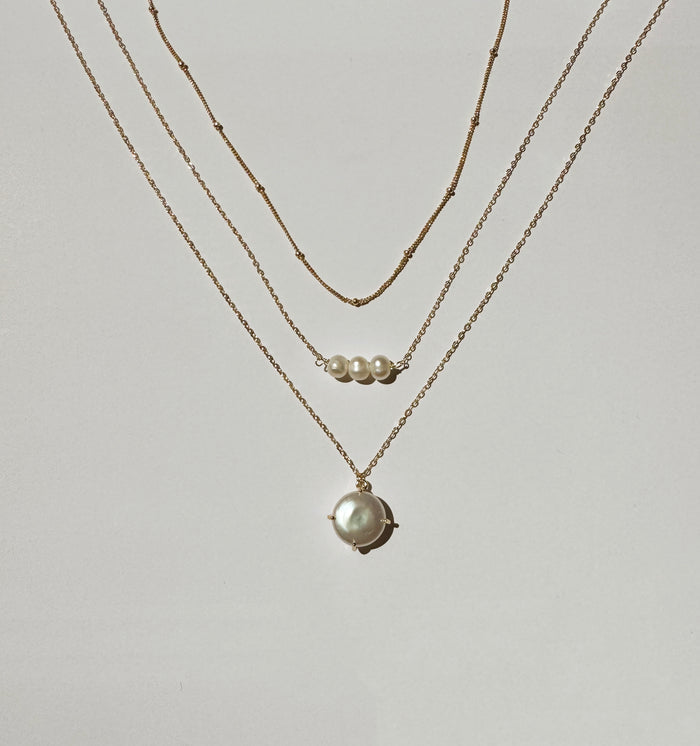 The Pearl Drop Set by LLUME Jewelry features three gold necklaces: two simple chains, one with three small pearls, and a stunning pearl pendant—all arranged on a white surface for a versatile, elegant look.