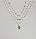 The Pearl Drop Set by LLUME Jewelry features three gold necklaces: two simple chains, one with three small pearls, and a stunning pearl pendant—all arranged on a white surface for a versatile, elegant look.