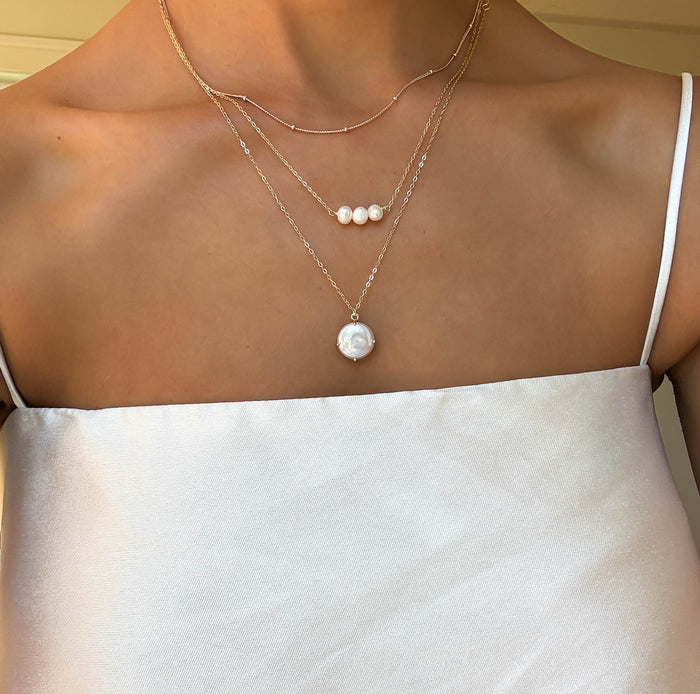 A woman in a white satin spaghetti strap top is shown from the shoulders down, wearing LLUME Jewelry’s Pearl Drop Set—layered gold necklaces adorned with delicate pearls and a bold pearl pendant.